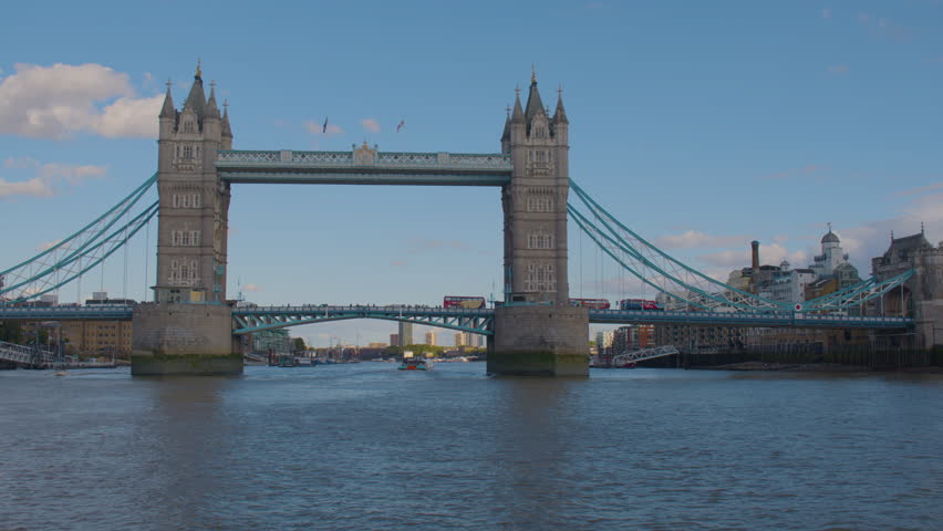 Tower Bridge spans the River Thames under a clear blue sky, with its twin Gothic towers, suspension cables, and passing traffic visible along the deck and boats moving below.