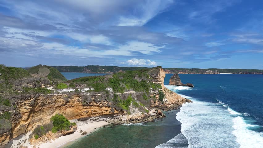 Aerial drone shot of stunning tropical coastline with dramatic cliffs, turquoise ocean waves crashing on white sandy beach, and picturesque rock formations under a vibrant blue sky.