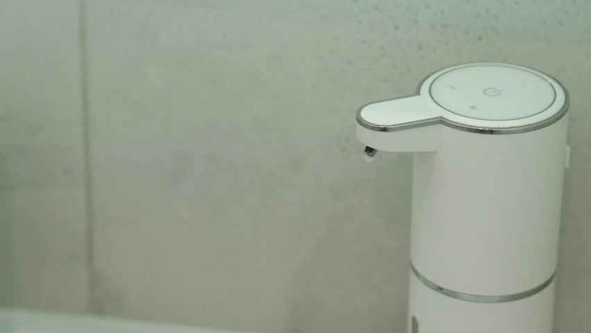 Hands approach a touchless dispenser which releases liquid soap in a bright bathroom. A quick and easy hand washing demonstration.