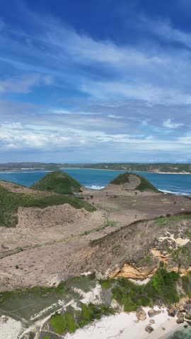 Aerial View of Beautiful Coastal Landscape with Lush Green Hills, Dry Fields, White Sandy Beach, Rocky Shore, and Calm Blue Ocean under a Bright Sky with Wispy Clouds