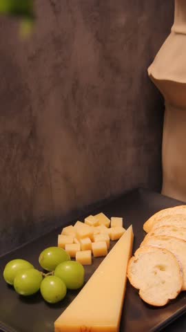 A vertical close-up of a parmesan, green grape, and bread platter on a black plate. The camera stays still with only a soft leaf movement at the top, creating a calm and elegant food scene.