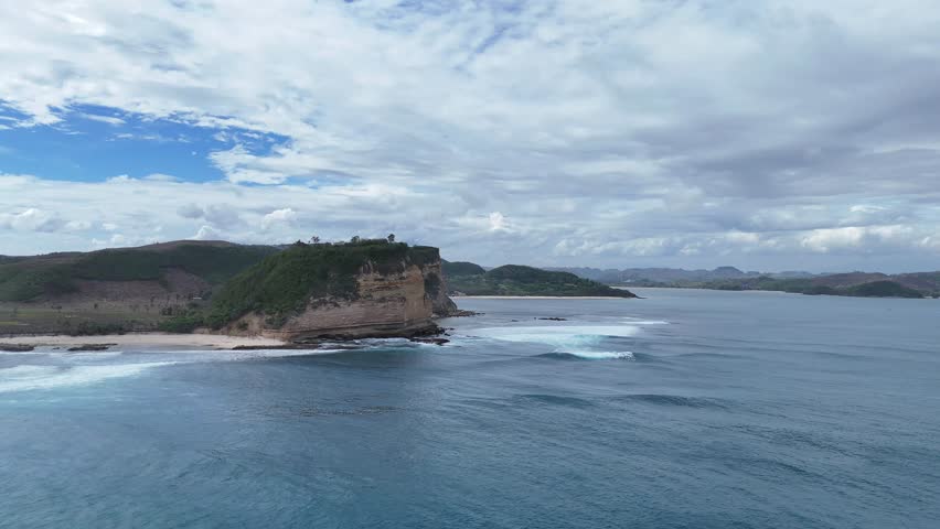 Aerial View of Dramatic Rocky Cliff Coastline with Lush Green Hills and Turquoise Ocean Waves Crashing on a Secluded Sandy Beach Under a Cloudy Sky