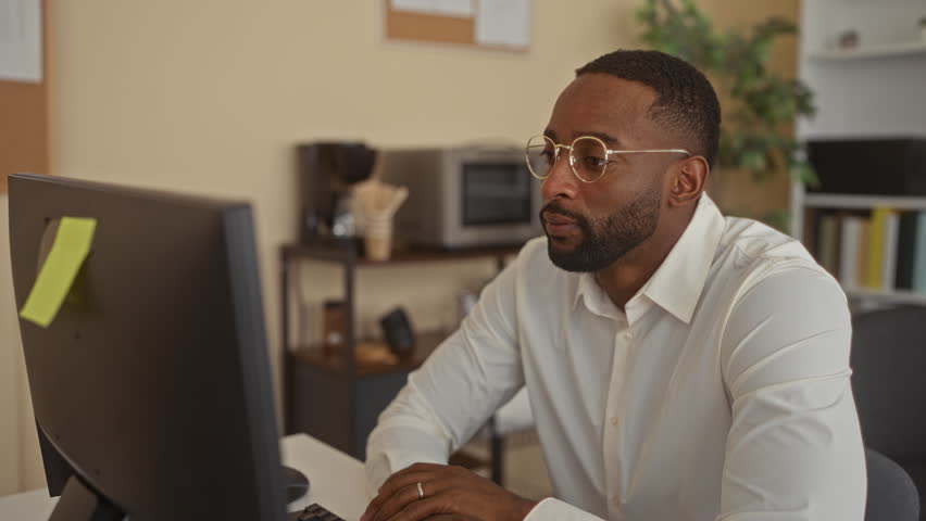 Man typing on keyboard with hands on keys, wearing round glasses and white shirt at a desk in an office building; focus productivity confidence.