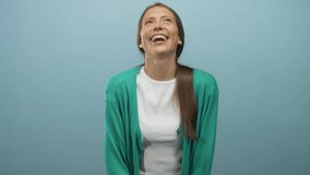 Young hispanic woman laughing with open mouth and tilted head in studio teal blue backdrop wearing green cardigan and white t shirt; joy. - Powered by Shutterstock - Get 15% off with code: PIKWIZARD15