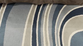 Close-up shot of a modern floor rug featuring wavy, blue and gray abstract stripes, meeting the plain dark brown bed skirt and white sheet above - Powered by Shutterstock - Get 15% off with code: PIKWIZARD15