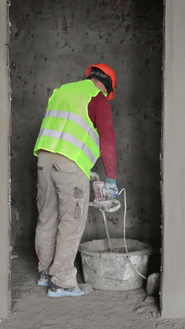 Construction worker mixing plaster in a bucket, Worker wearing safety gear stirring plaster with a mixer.