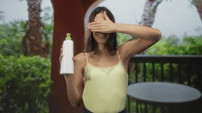 Woman holding white pump bottle and hand covering eyes on building balcony with palm trees and round table visible; playful skincare. - Powered by Shutterstock - Get 15% off with code: PIKWIZARD15