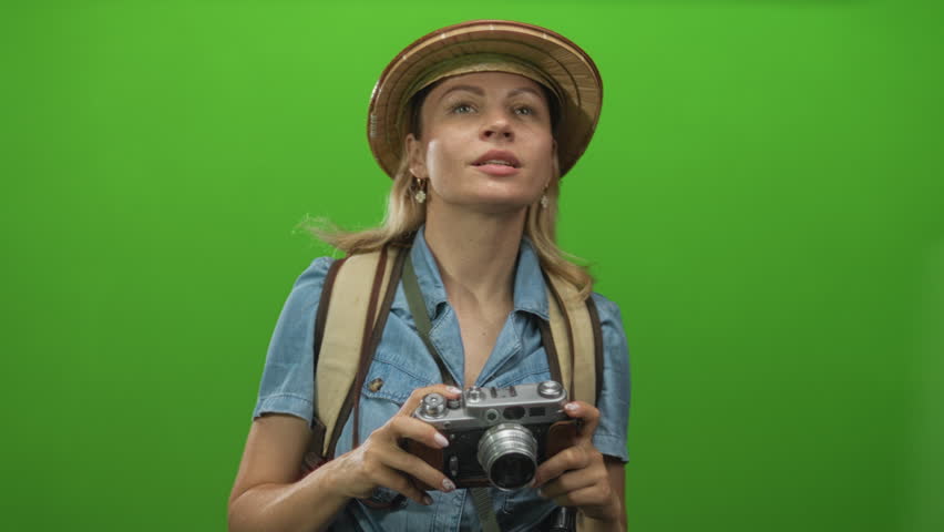 Blonde explorer woman in straw hat raising camera with both hands and gripping backpack strap in studio; curiosity.