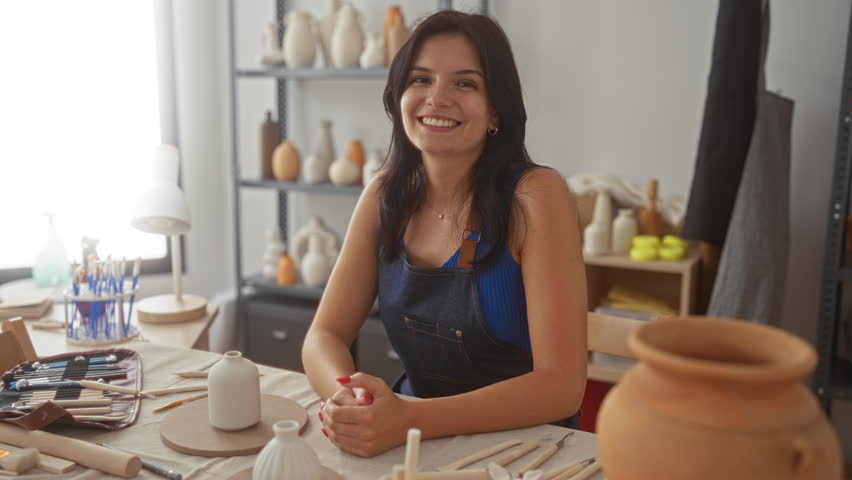 Woman with ceramic vase and hands clasped at pottery worktable in studio; serenity creativity passion craftsmanship inspiration imagination innovation artistry vision expression.