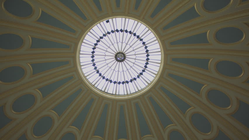 Tilt shot of a grand circular library interior with high arched windows, domed ceiling, and towering wooden bookshelves filled with books, illuminated by soft natural light and classic reading lamps.