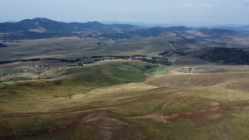 Aerial footage of a vast mountain range with rolling green hills. A dirt road snakes along the terrain, leading towards a distant valley