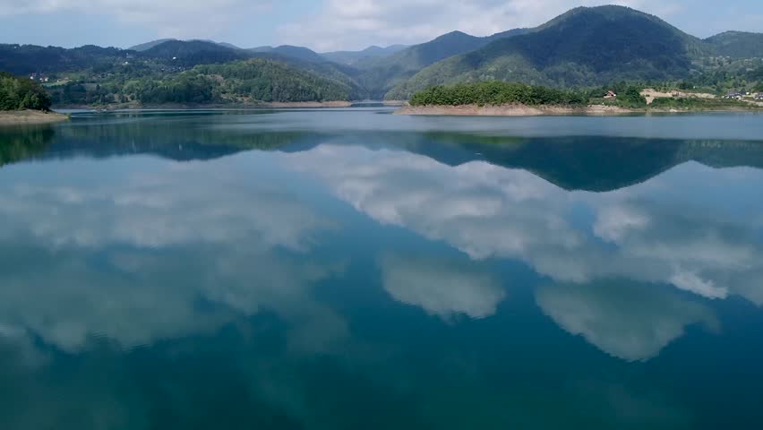 Aerial footage of a calm lake Zaovine surface reflecting the blue sky and white clouds. Beautiful natural landscape with green mountains and hills