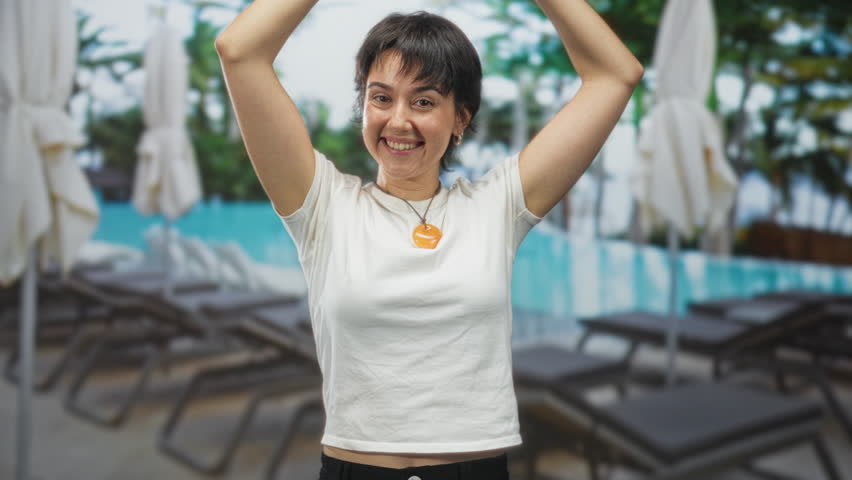 Woman with orange pendant necklace, arms raised and smiling while standing poolside at a building; joy celebration.