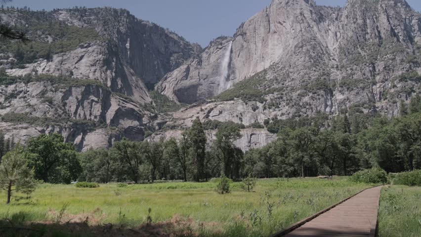 Upper Yosemite Falls Viewed from Meadow Boardwalk with Granite Cliffs
A wooden boardwalk through a bright Yosemite Valley meadow towering granite cliffs and Upper Yosemite Falls, framed by trees