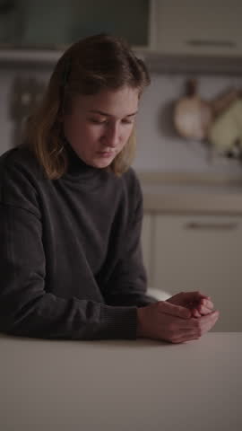 Vertical portrait of young Muslim woman with cerebral palsy sitting at table, looking down and clasping hands in moment of quiet prayer and thoughtful reflection in supportive home environment.