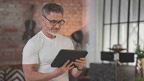 Middle aged man using digital tablet at home in loft living room. Happy, sporty, active senior male with gray hair and glasses reading social media, working and smiling. - Powered by Shutterstock - Get 15% off with code: PIKWIZARD15