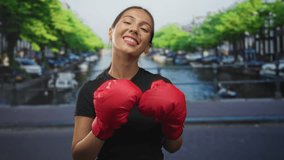 Woman with red boxing gloves smiling with closed eyes and raised fists on street by amsterdam canal bridge; confidence motivation. - Powered by Shutterstock - Get 15% off with code: PIKWIZARD15