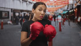 Woman wearing black tee holds red boxing gloves up in guard, fists raised on busy city street lined with lanterns; confidence resilience strength. - Powered by Shutterstock - Get 15% off with code: PIKWIZARD15