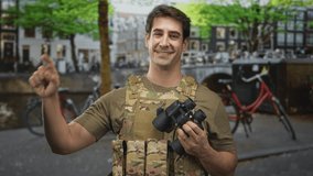 Man soldier in camouflage vest holds binoculars and points right index finger up on a street canal bridge; duty pride. - Powered by Shutterstock - Get 15% off with code: PIKWIZARD15