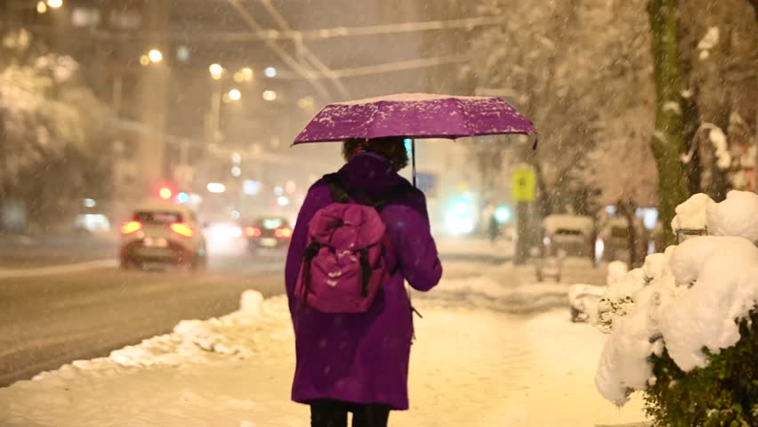 A woman with an umbrella walks in the city while it is snowing in winter. Snowflakes falling in the city streets. Snowy night.