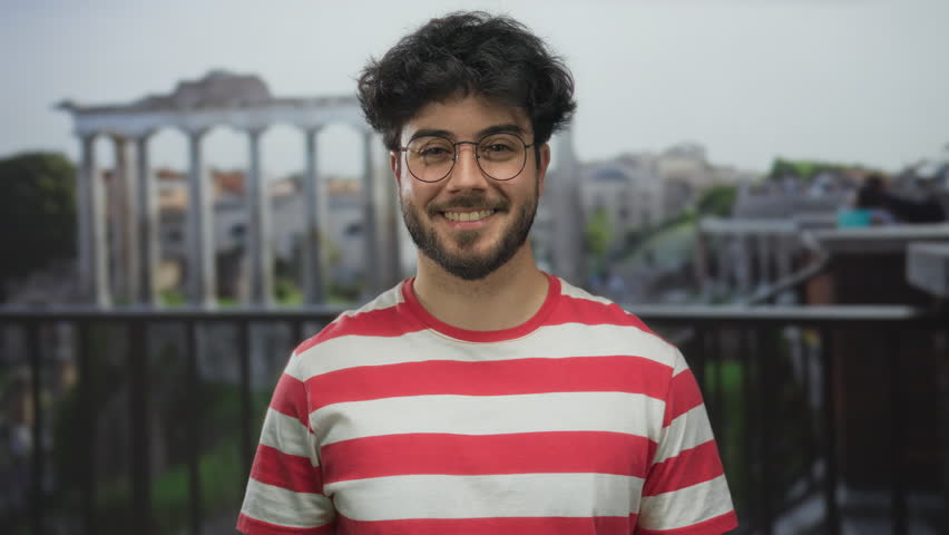 Man with beard and glasses in red striped shirt smiling outdoors at roman ruins on a sunny day.