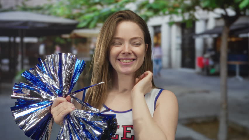 Young cheerleader woman holding blue metallic pompom points finger and smiles widely on street; enthusiasm.