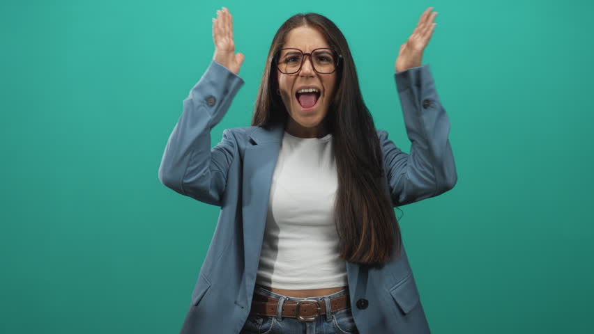 Young woman raises hands to head, mouth open and wearing glasses with visible hands and belt in teal studio; surprise.