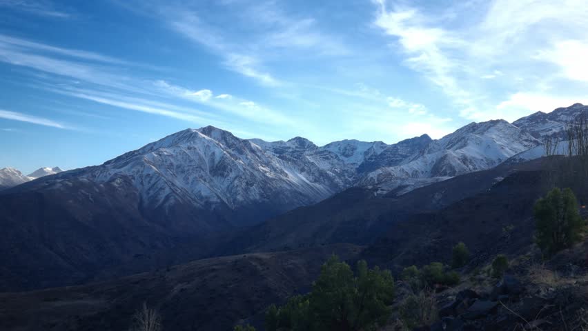 Landscape image showing the snow-covered mountains of Andes Mountains beneath a blue sky with thin clouds, in Chile.	