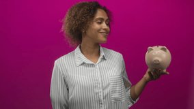 Woman holding piggy bank with palm up and smiling, wearing striped shirt in magenta studio; savings budgeting confidence. - Powered by Shutterstock - Get 15% off with code: PIKWIZARD15