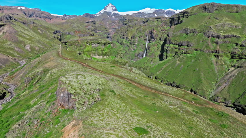 Aerial view of open valley below Mulagljufur Canyon. Drone footage showing grassy plains leading toward tall green cliffs with waterfalls in the distance.