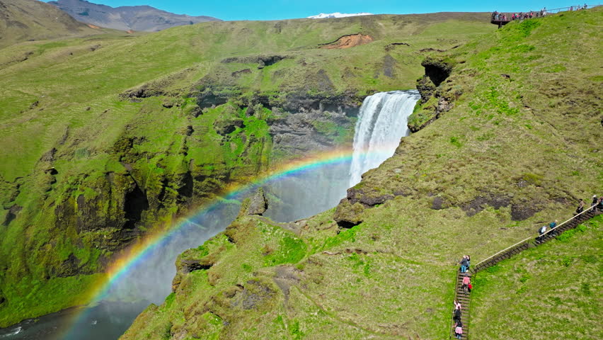Aerial view of Skogafoss waterfall from the canyon edge. Drone footage showing mist rising from the falls and a bright rainbow curving across the rugged landscape.