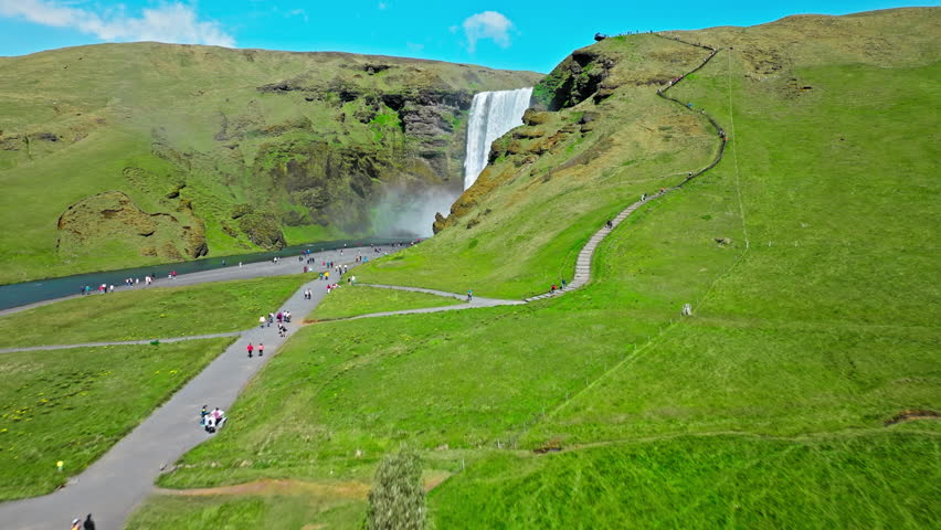 Aerial view of Skogafoss waterfall in bright sunlight. Drone footage showing a clear rainbow forming in the mist as water plunges over the cliff into the valley.