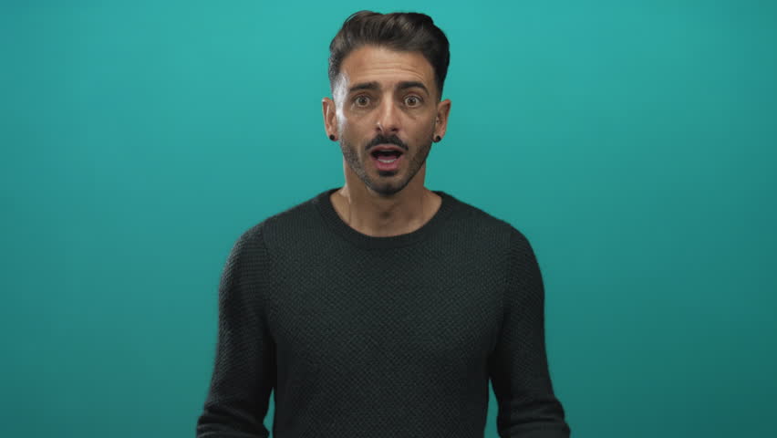 Man pointing finger to chest and shrugging hands with open mouth and clenched fist pump in teal studio; surprise.