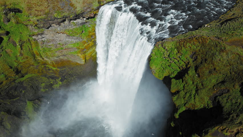 Close aerial view of Skogafoss waterfall from above. Drone footage highlighting rushing water and mist swirling upward as light catches rainbow hues.