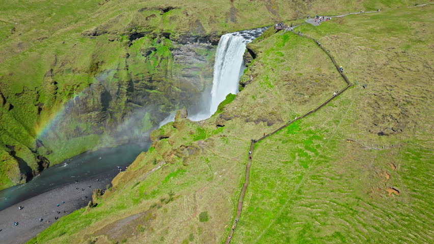 Aerial view of Skogafoss waterfall dropping over a rugged cliff. Drone footage showing white mist rising from the canyon and sunlight revealing subtle rainbow colors.