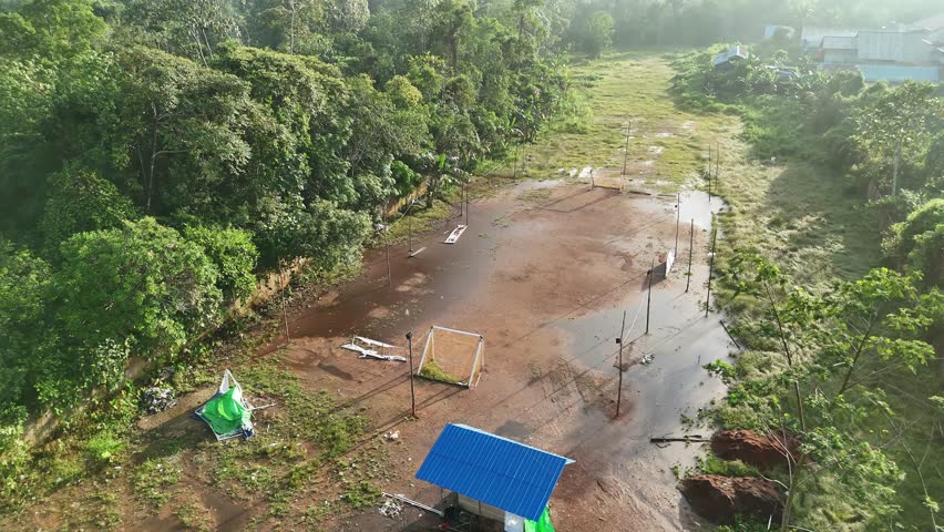 An aerial view of a simple football field covered in mud and partially flooded with water, equipped with small goals and benches, surrounded by lush trees.