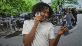 Woman holds smartphone to ear and blue creditcard on sunlit street lined with bicycles and leafy trees; satisfaction. - Powered by Shutterstock - Get 15% off with code: PIKWIZARD15