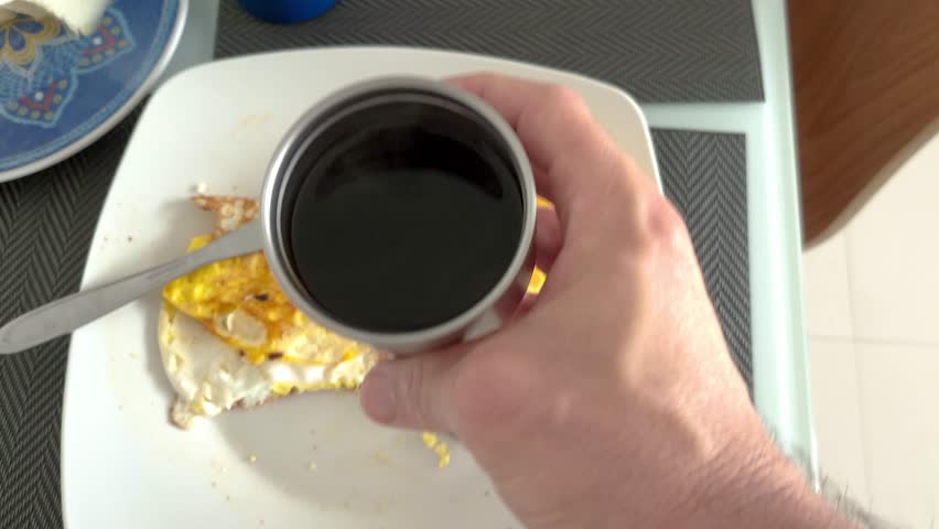 Man holding a steaming cup of coffee over a plate with fried eggs.