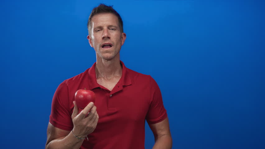 Man wearing red polo shirt holding red apple in one hand and resting other hand on chest with slight smile in blue studio; temptation.