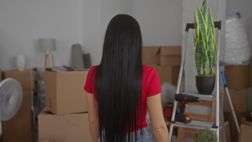 Woman standing in a new home surrounded by moving boxes, tools, fan, and ladder, wearing a red shirt, in a cluttered interior apartment setting with a houseplant.