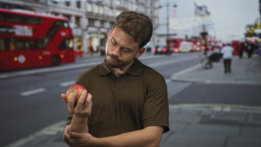 Man holding apple with hand on chin on busy city street, red bus blurred behind him and pedestrians on the sidewalk; thoughtful.