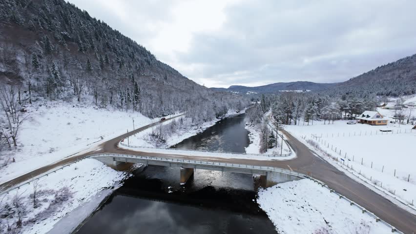 Cinematic aerial view of a winter river winding between snowy trees and mountain slopes. Cold dramatic atmosphere captured with FPV drone for nature and landscape projects.
