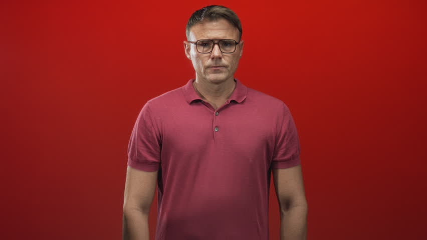 Man with glasses in a maroon polo pressing his hand to temple and closing his eyes against a red studio backdrop; stress headache.