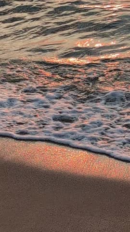Close-up shot captures the rhythmic motion of gentle ocean waves rolling onto the sandy shore during the golden hour.