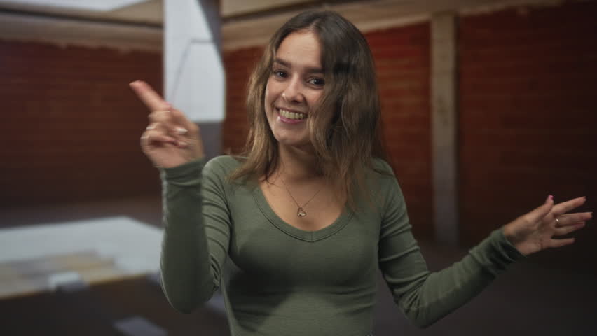 Woman smiling with hands out in building, casual green top and heart pendant, motion blur on hands and relaxed pose; optimism warmth joy.