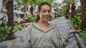 Woman smiling and holding palm up in gray hoodie on street promenade showing open hand gesture; happy friendly welcome. - Powered by Shutterstock - Get 15% off with code: PIKWIZARD15