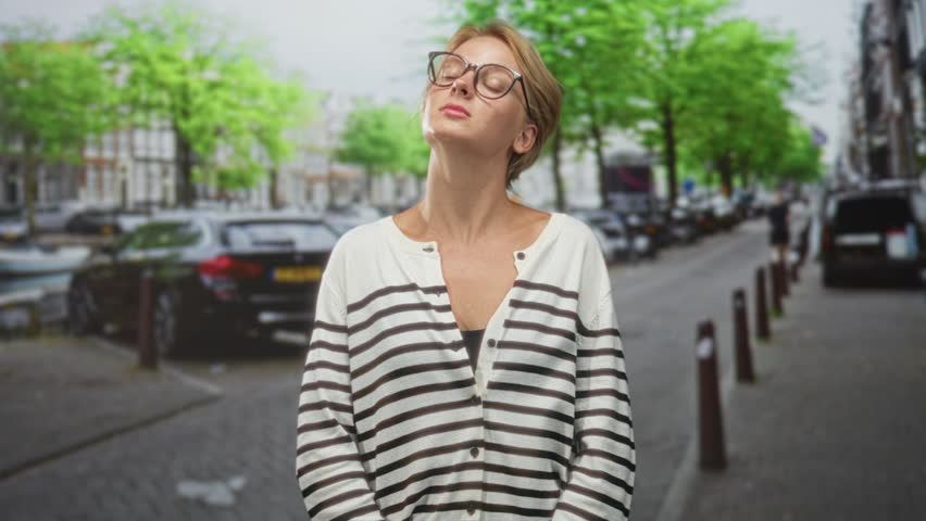 Blond woman holding car keys and raising palm in stopping gesture on street, wearing striped cardigan and glasses, pressing key fob with other hand; hesitation privacy. - Powered by Shutterstock - Get 15% off with code: PIKWIZARD15
