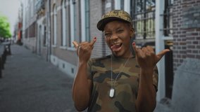 Woman soldier wearing camouflage cap and uniform sticks out tongue and makes rock horns gesture on a street; fun. - Powered by Shutterstock - Get 15% off with code: PIKWIZARD15