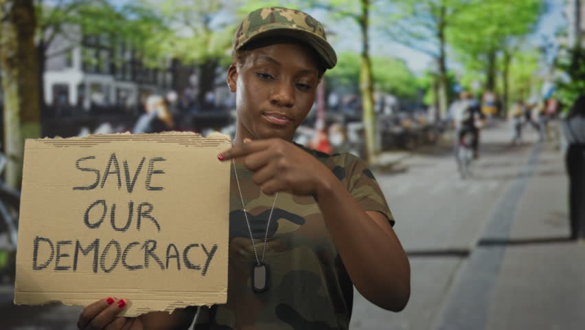 Young african american woman in camouflage uniform holds cardboard sign reading save our democracy on busy street; activism.