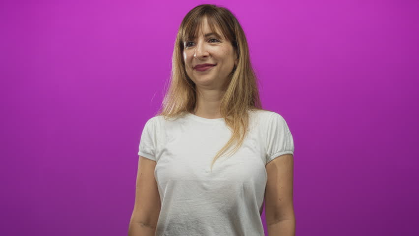 Woman smiling wearing white t shirt and hoop earring with head tilt in purple studio; joy warm friendly.